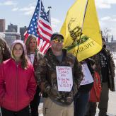 Small group poses for camera with US and Gadsden flags. In front, a child in pink jacket stands beside man wearing a brown camo jacket with sign pinned on it. Sign reads: "GUN FREE ZONES ARE KILLING OUR KIDS"