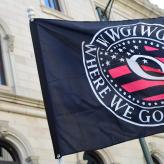 A big black flag that has a Q on the middle with red and black stripes and the words Where We Go One We Go All around it.