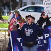A White woman with blond hair, wearing a hat. She has a Trump 2020 sweatshirt on, and is holding an Amerian Flag and a water bottle. Her mouth is open like she's shouting something.