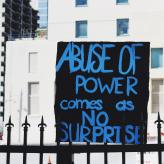 A black sign that says "Abuse of power comes as no surprise."