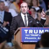A man in a suit with a receding hairline talking into a microphone.