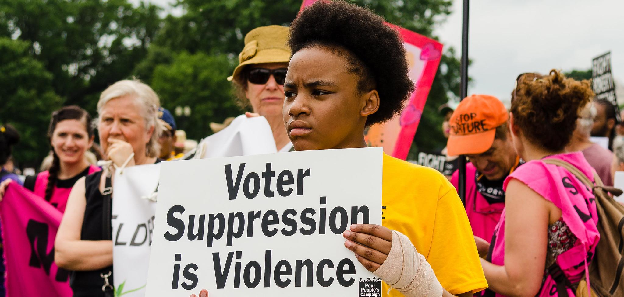 A Black person in a yellow T-shirt holding a sign that says "voter suppression is violence"